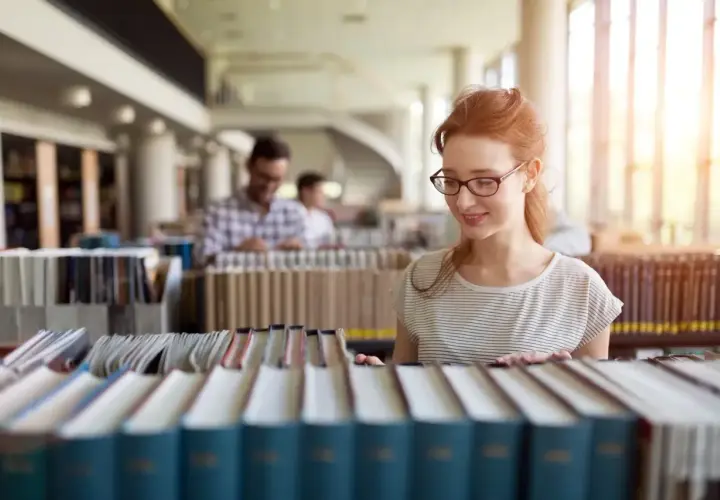 Studentka z rudymi włosami w okularach przegląda książki w bibliotece, szukając materiałów do prawo studia.