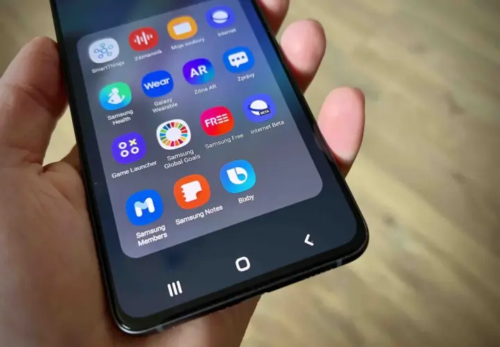 Dłoń trzyma smartfon z widocznym folderem aplikacji, w tym ikoną Bixby.