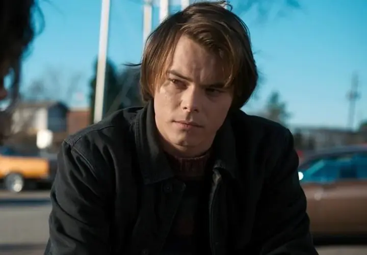 Charlie Heaton w ciemnej kurtce i swetrze, zamyślony, na tle błękitnego nieba i samochodów.