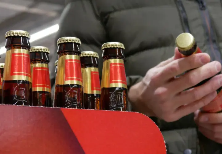 Rząd butelek piwa w czerwono-złotych etykietach. Człowiek w zielonej kurtce trzyma jedną butelkę, zastanawiając się, ile kosztuje jedno piwo.