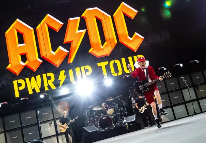AC/DC na koncercie w Polsce. Wokalista w czerwonym stroju skacze z gitarą na tle gigantycznego logo zespołu i ściany wzmacniaczy.