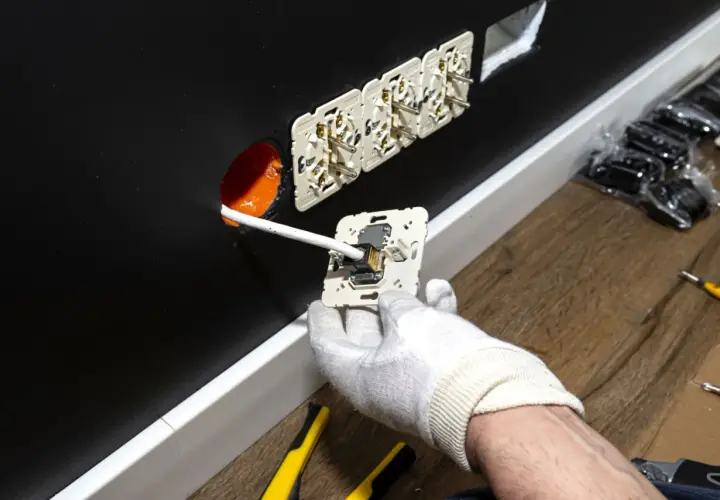 Instalacja gniazdek elektrycznych. Ręka w białej rękawiczce podłącza przewód do mechanizmu gniazdka. W ścianie widać już kilka zamontowanych gniazdek. Zastanawiasz się, ile za punkt elektryczny?