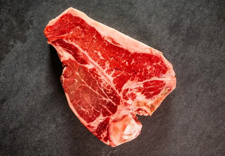 Una succulenta bistecca di manzo T-bone, con la sua marmorizzazione di grasso, pronta per essere cucinata su una superficie scura.