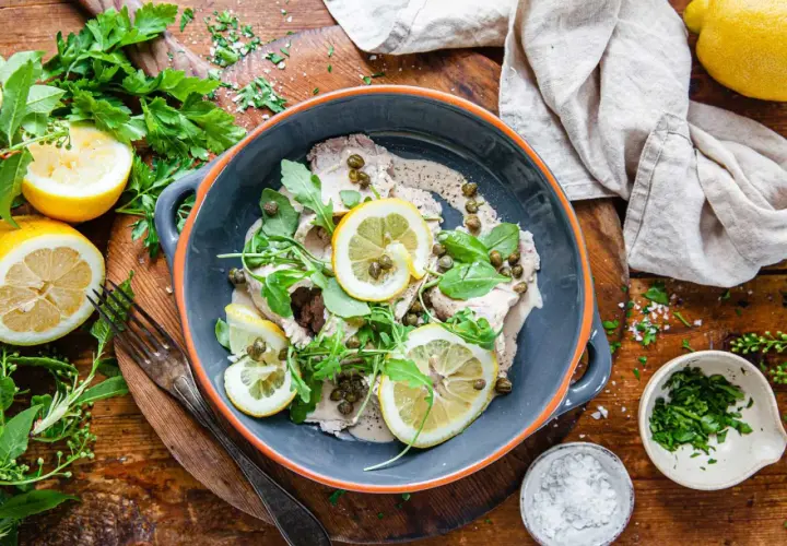 Vitello tonnato i en grå skål, garnerad med citronskivor, kapris och ruccola. Serveras med färska örter och citroner på ett träbord.