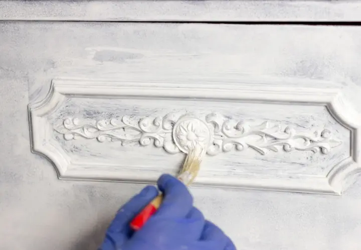 Mano con guanto blu che applica chalk paint fai da te su un mobile con decorazioni in rilievo, creando un effetto shabby chic.