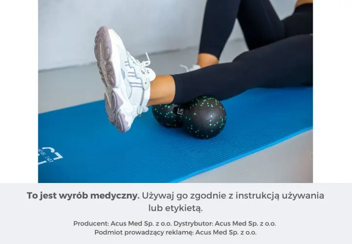 Kobieta wykonuje rolowanie mięśni łydki za pomocą czarnego rollera z zielonymi kropkami, leżąc na niebieskiej macie.