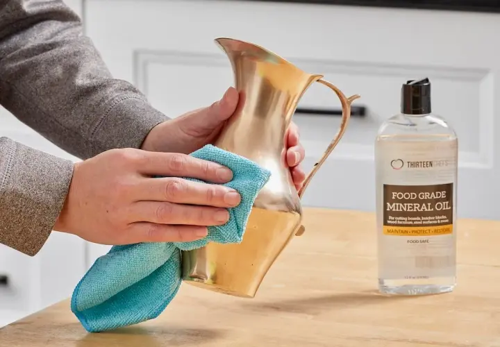 Hands polish a brass pitcher with a blue cloth, showing how to clean brass with mineral oil.
