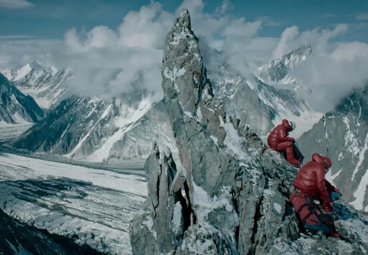 Dwóch alpinistów w czerwonych kurtkach odpoczywa na skalistej grani, podziwiając lodowiec i ośnieżone szczyty. Widok na berbeka broad peak zapiera dech w piersiach.