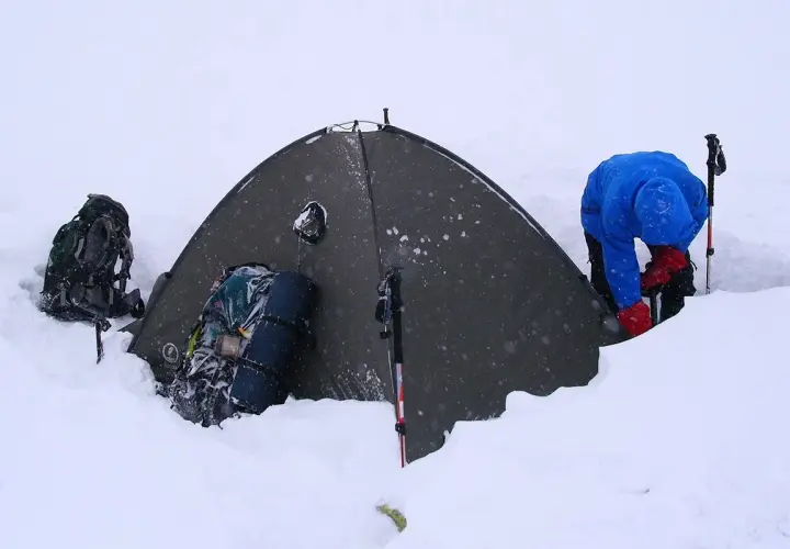 Osoba w niebieskiej kurtce i czerwonych rękawiczkach przygotowuje namiot w śniegu. Obok leżą plecaki i kijki trekkingowe, które pomagają dobrać kijki trekkingowe do wzrostu.