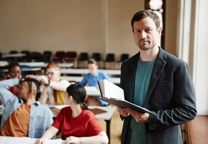 Nauczyciel z książką w ręku, przygotowujący się do lekcji. W tle widać uczniów, co sugeruje jego pensum nauczyciela.