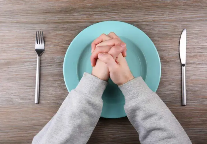 Manos entrelazadas sobre plato vacío, tenedor y cuchillo a los lados. Quizás piensa que no cenar adelgaza.