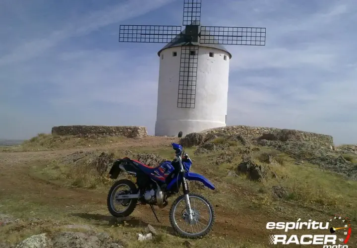 Yamaha DT 125 RE azul y roja aparcada junto a un molino de viento blanco en un paisaje rocoso.