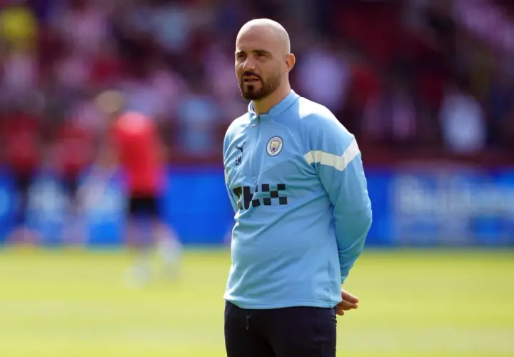 Łysy mężczyzna z brodą, trener Manchester City, w błękitnej bluzie treningowej, stoi na boisku.