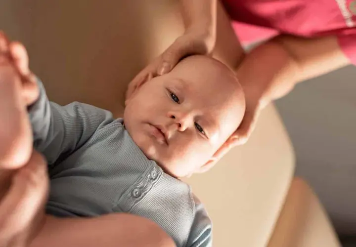 Niemowlę w niebieskim body, któremu fizjoterapeuta delikatnie masuje główkę.