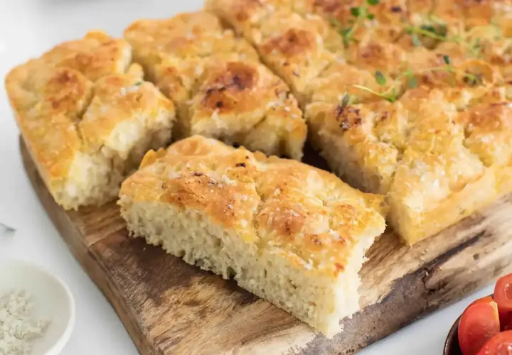 Focaccia dorada y tierna con sal gruesa y romero, cortada en porciones sobre tabla de madera.