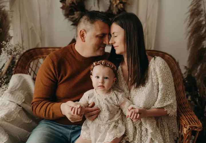 Ciepła sesja na roczek z rodzicami. Malutka córeczka w koronkowej sukience trzyma dłonie mamy i taty, którzy patrzą na nią z miłością.
