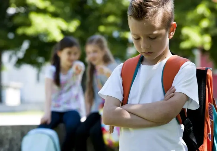 Ein Junge mit Rucksack steht traurig da, während zwei Mädchen ihn im Hintergrund auslachen. Ein trauriges Bild von Mobbing in der Grundschule.