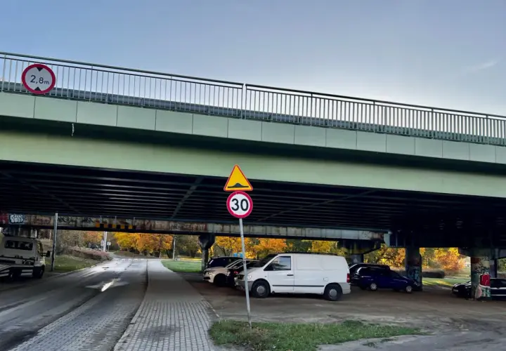 Pod estakadą zaparkowane są samochody. Widoczny znak ograniczenia wysokości 2,8m i znak ograniczenia prędkości do 30 km/h.