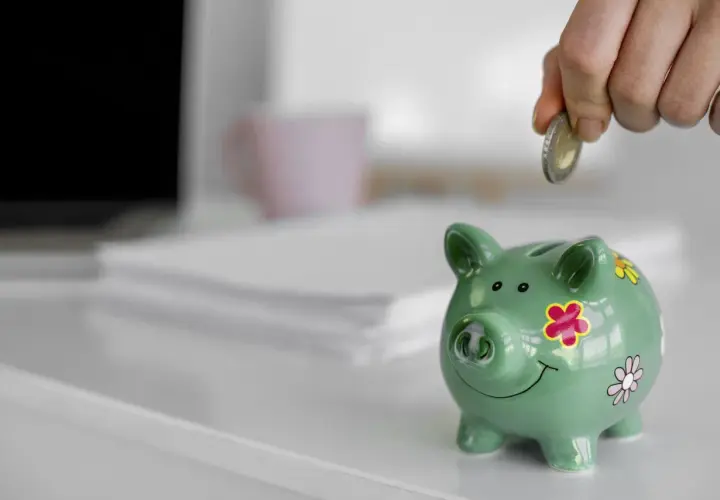 Ręka wrzuca monetę do zielonej świnki skarbonki. To symbol oszczędzania i finansowego zabezpieczenia.