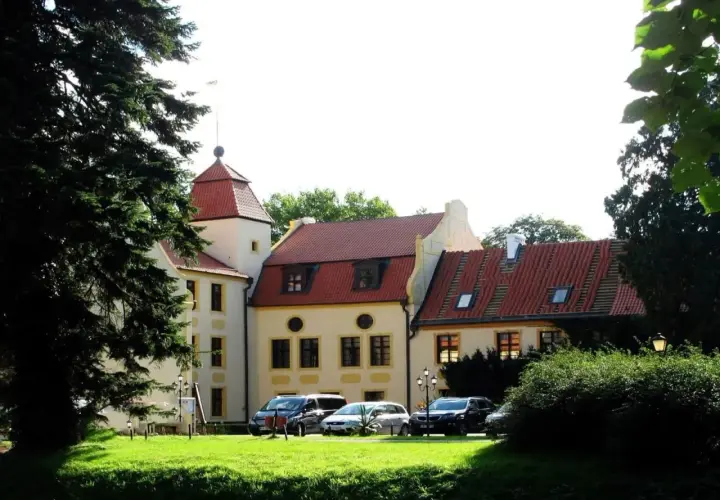 Pałac w Krokowej z czerwoną wieżą i dachami, otoczony zielenią. Przed budynkiem zaparkowane samochody.