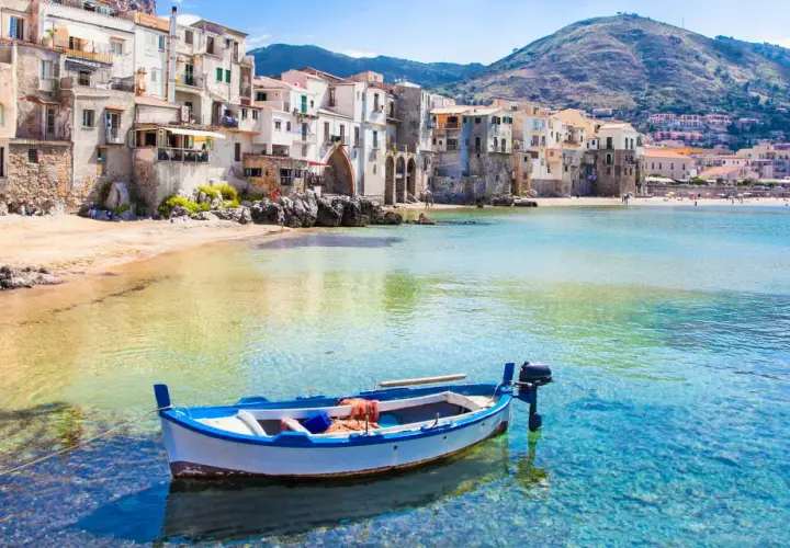 Bateau blanc et bleu ancré dans les eaux cristallines de la plage de Cefalù, avec des maisons colorées en arrière-plan.