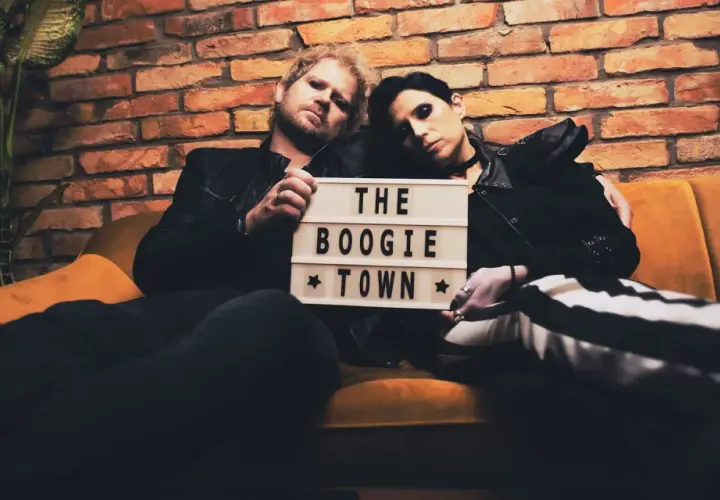 Para siedzi na kanapie, trzymając tabliczkę z napisem "THE BOOGIE TOWN".