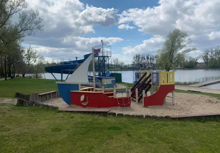 Kolorowy plac zabaw w kształcie statku na piaszczystej plaży nad jeziorem Średzkim. W tle widać zjeżdżalnie wodne i spokojną taflę wody.