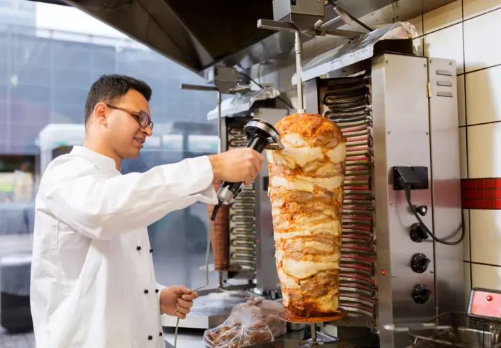 Kucharz w białej kurtce kroi mięso z pionowego rożna na kebab.