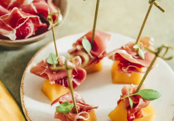 Une présentation originale de melon et jambon, des brochettes savoureuses sur une assiette blanche, avec un melon entier sur le côté.