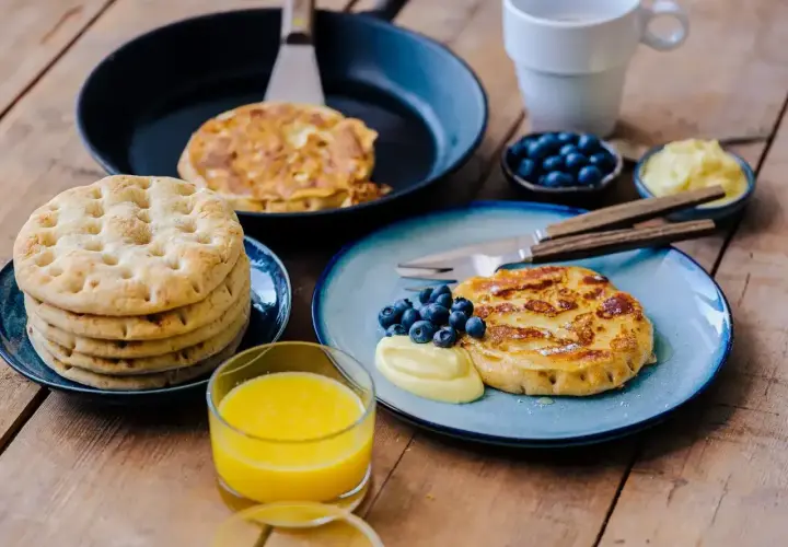 Fattiga riddare med blåbär och vaniljsås på en träyta. En stapel tunna pannkakor och ett glas apelsinjuice.