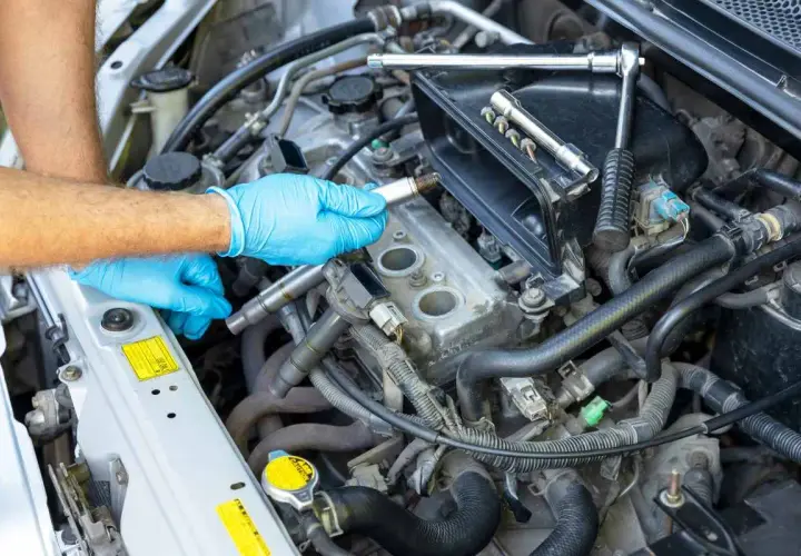 Mano con guante azul sosteniendo una bujía, lista para el cambio de bujías en el motor de un coche. Herramientas a la vista.