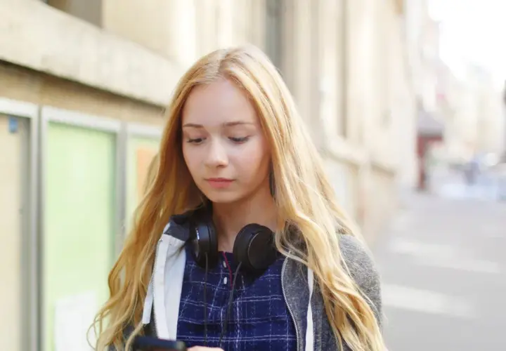 Dziewczyna z blond włosami i słuchawkami na szyi przegląda telefon, zastanawiając się, jak zacząć rozmowę z chłopakiem.