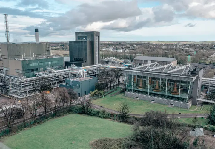 Vue aérienne de la distillerie Midleton, avec ses bâtiments industriels, ses cuves en cuivre visibles à travers une façade vitrée, et un paysage verdoyant en arrière-plan.