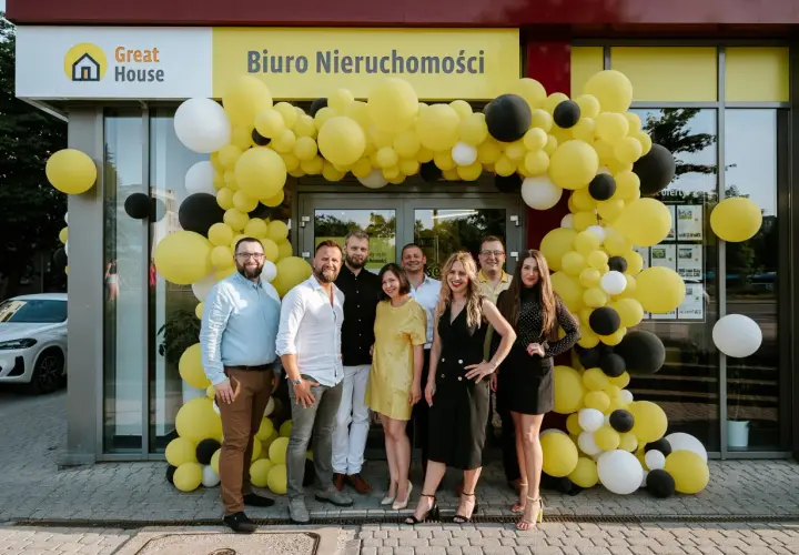 Grupa ludzi pozuje przed biurem nieruchomości ozdobionym żółtymi i czarnymi balonami.