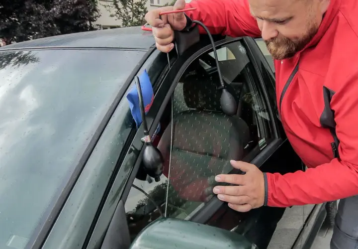 Mężczyzna w czerwonej kurtce wykonuje otwieranie awaryjne samochodu, używając narzędzia do podważenia drzwi.