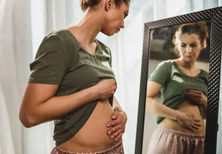 Mujer se mira al espejo, tocándose el abdomen con preocupación por la hinchazón después de comer.