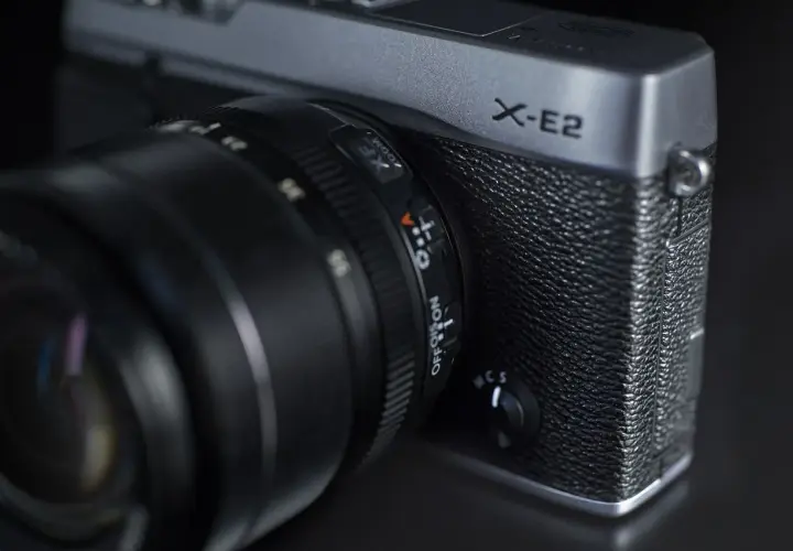 Srebrny aparat Fujifilm X-E2 z obiektywem. Widoczny napis "X-E2" i pokrętło wyboru trybu.