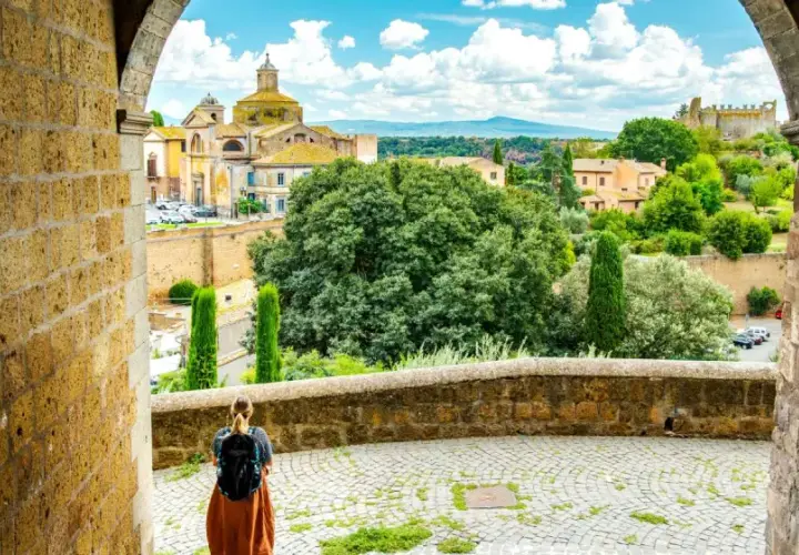 A traveler pauses, backpack ready, gazing at the historic Italian landscape. This view, perhaps on a journey from Viterbo to Rome, showcases ancient architecture and lush greenery under a bright sky.