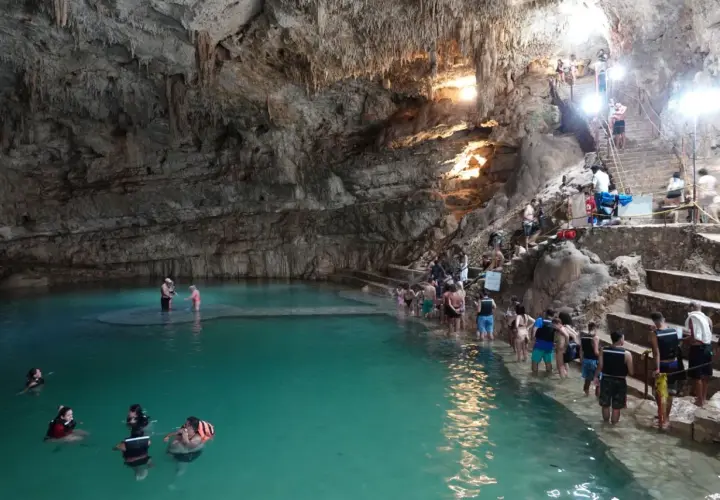 Menschen schwimmen und stehen im türkisfarbenen Wasser einer Höhle in Valladolid, Mexiko. Treppen führen zu einer Plattform, auf der sich weitere Besucher aufhalten.