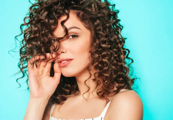 Mujer con cabello castaño y rizos definidos, tocando su cabello con la mano, sobre un fondo azul turquesa.