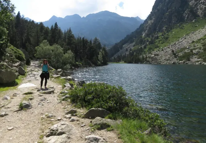 Una mujer camina por un sendero rocoso junto a un estany llong, rodeado de montañas y pinos.