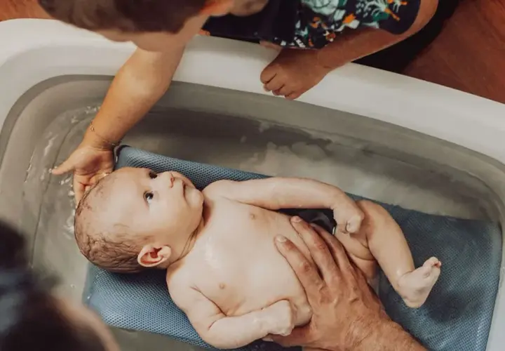 Delikatna kąpiel noworodka w wanience. Starsze dziecko z zaciekawieniem obserwuje maluszka.