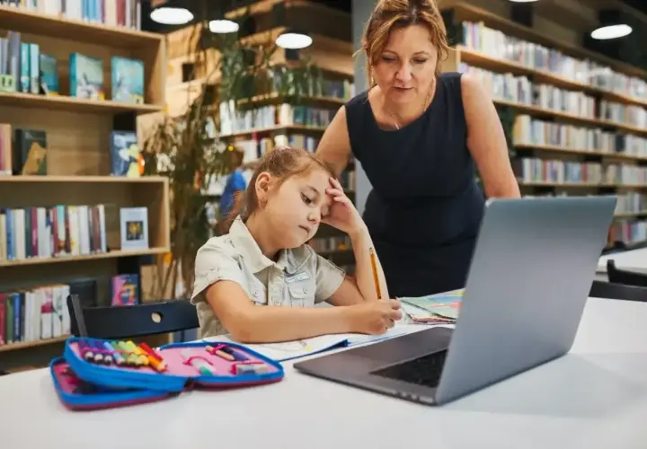 Dziewczynka z ołówkiem w dłoni, zamyślona nad zadaniem, przygotowuje podanie do szkoły średniej. Nauczycielka wspiera ją w bibliotece.