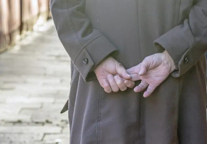 Osoba w brązowym płaszczu, z rękami splecionymi za plecami, stoi na chodniku.