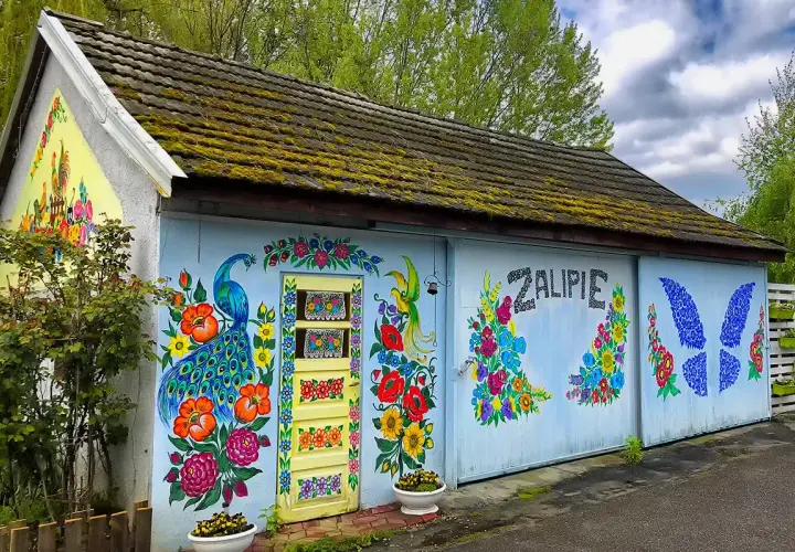 Kolorowy dom w Zalipiu ozdobiony malowidłami kwiatów i pawia. Na ścianie widnieje napis "Zalipie cennik".