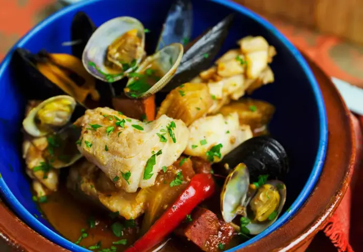Delicioso bacalao con almejas y gambas, servido en un cuenco azul con perejil picado y un chile rojo.