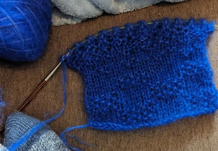 Un échantillon de tricot bleu vif, avec une pelote de laine assortie et une aiguille. La jauge maille est bien visible sur le tissu.
