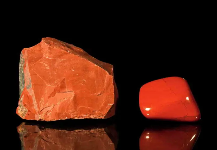 Zwei rote Jaspis-Arten: ein roher Stein und ein polierter Trommelstein, die auf schwarzem Hintergrund reflektieren.