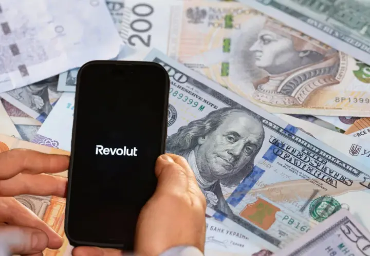 Telefon z logo Revolut na tle różnych walut. Może to symbolizować globalne finanse lub potencjalne zajęcie komornicze w kontekście międzynarodowych transakcji.
