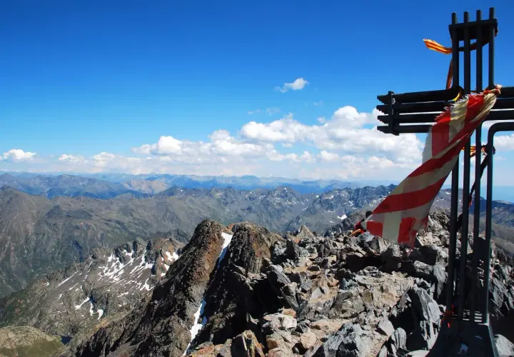 Cruz metálica en la cima de una pica d'estats, con bandera ondeando y un paisaje montañoso al fondo.